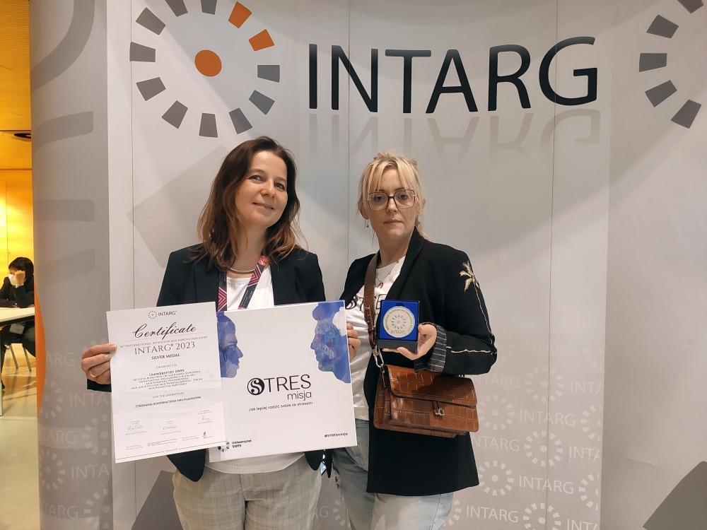 Dwie kobiety na tle ścianki z napisem „INTARG”. Kobieta po lewej trzyma certyfikat, kobieta po prawej – srebrny medal.