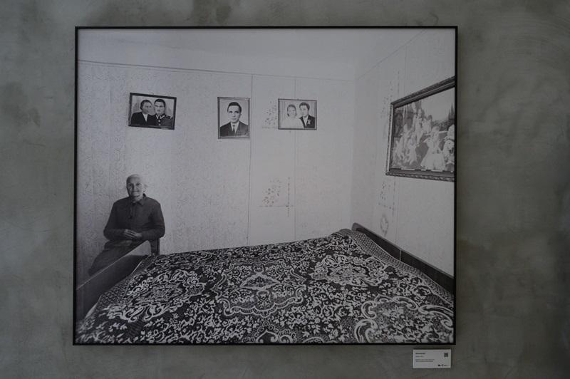 Fotografia autorstwa Zofii Rydet wyeksponowana na ścianie w czarnej ramie. Na zdjęciu widać starszą kobietę siedzącą w pokoju, obok dużego łóżka z wzorzystą narzutą. Na ścianach pokoju wiszą zdjęcia.
