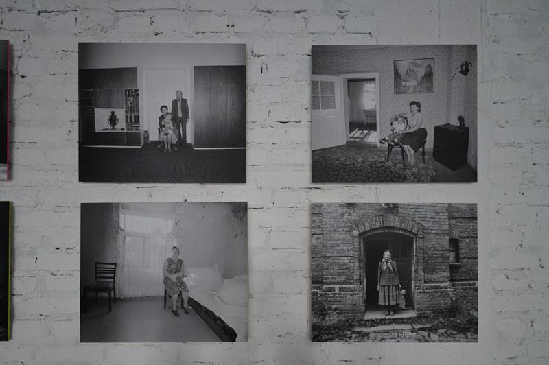 Cztery fotografie autorstwa Zofii Rydet wyeksponowane na ścianie. Na pierwszym zdjęciu od góry widać trzyosobową rodzinę: tatę i mamę z małym dzieckiem na kolanach, którzy znajdują się na środku pokoju z masywną komodą i dużym regałem. Na drugim zdjęciu widać młodą, uśmiechniętą kobietę siedzącą w salonie na krześle. Trzecie zdjęcie przedstawia kobietę siedzącą na krześle postawionym koło łóżka. Na czwartym zdjęciu widać starszą kobietę stojącą w środku ceglanej bramy wejściowej.  