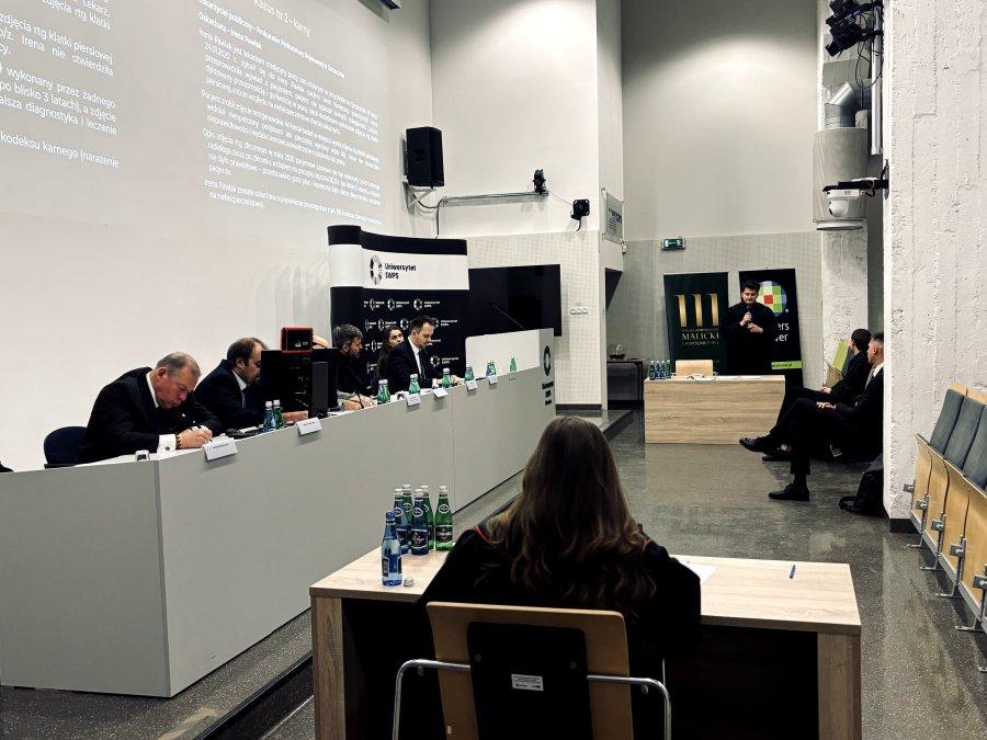 Student wygłasza mowę, jury robi notatki, kobieta w todze prokuratorskiej uważnie słucha