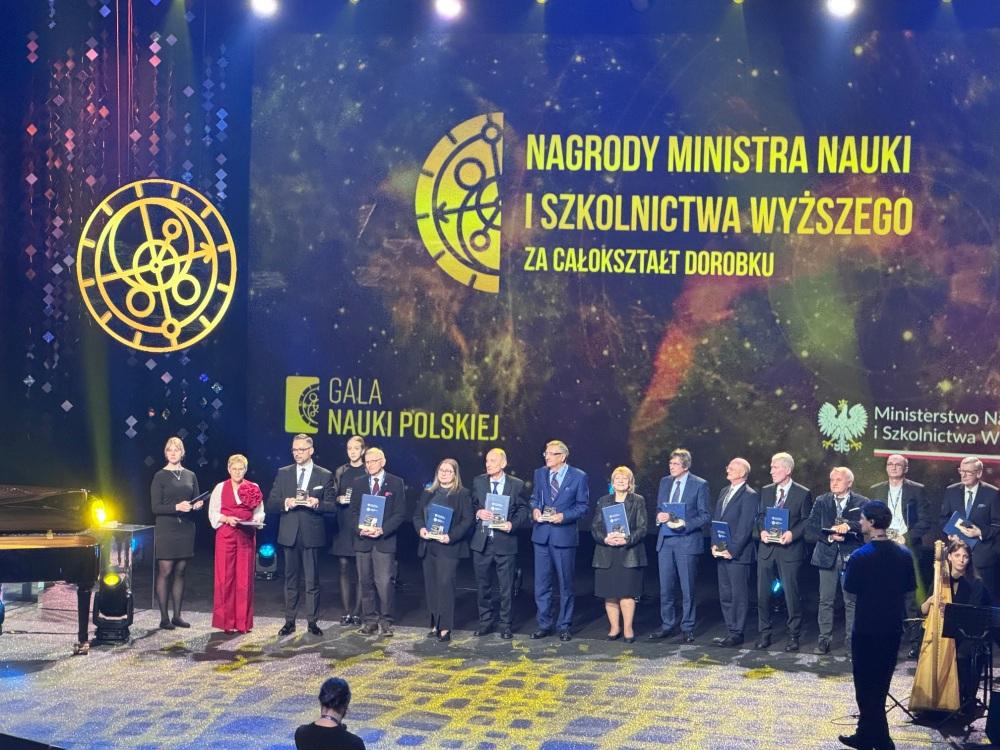 Kilkanaście elegancko ubranych osób stoi na scenie, trzymając nagrody. W tle napis „Nagrdy Ministra Nauki i Szkolnictwa Wyższego za całokształt dorobku. Gala Nauki Polskiej